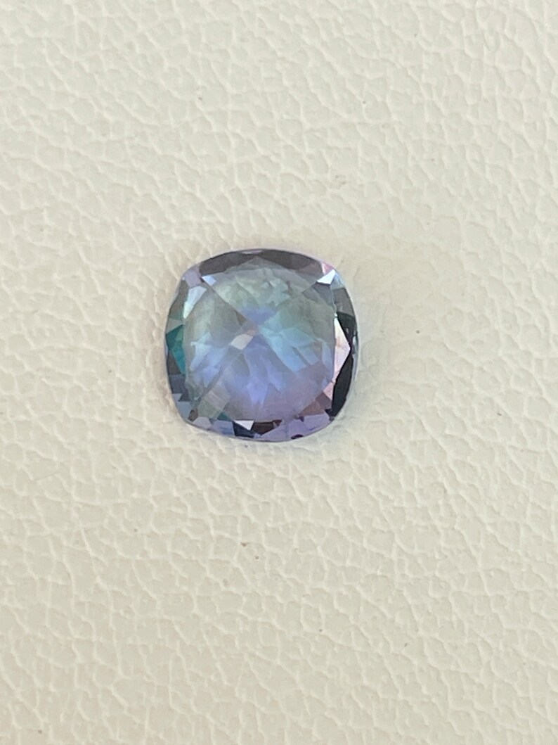 May include: A cushion-cut gemstone with a purple and blue hue. The gemstone is set against a white background.