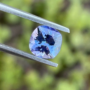 May include: A cushion-cut, purple gemstone with a light blue hue, held by tweezers against a blurred green background.