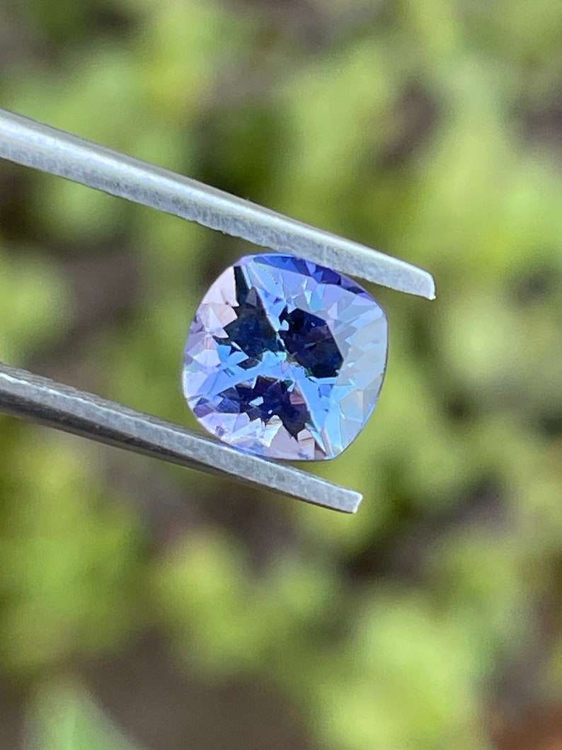 May include: A cushion-cut, purple gemstone with a light blue hue, held by tweezers against a blurred green background.