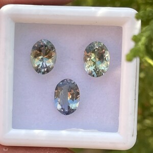 May include: Three oval-shaped gemstones in a white box. The gemstones are a light green, a darker green, and a light blue.