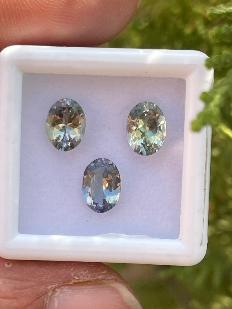 May include: Three oval-shaped gemstones in a white box. The gemstones are a light green, a darker green, and a light blue.