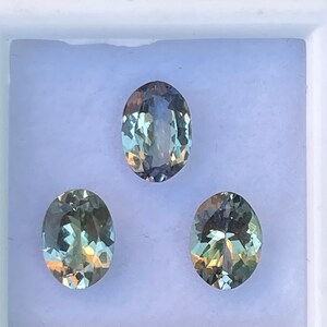 May include: Three oval-shaped gemstones in a white box. The gemstones are a light green color with a slight blue hue.