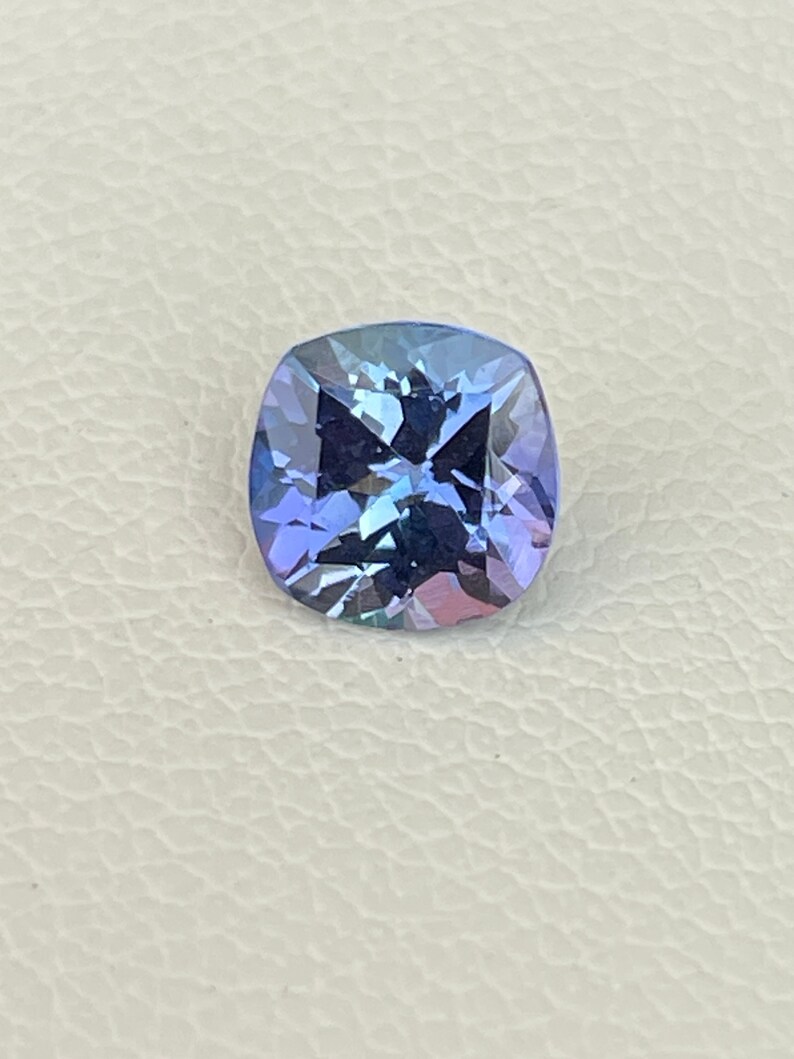 May include: A cushion-cut gemstone with a deep blue and purple hue. The gem is faceted and has a high level of brilliance.