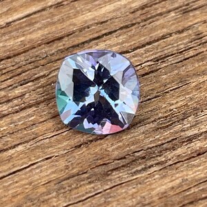 May include: A cushion-cut gemstone with a purple hue and iridescent rainbow reflections. The gem is set against a wooden background.