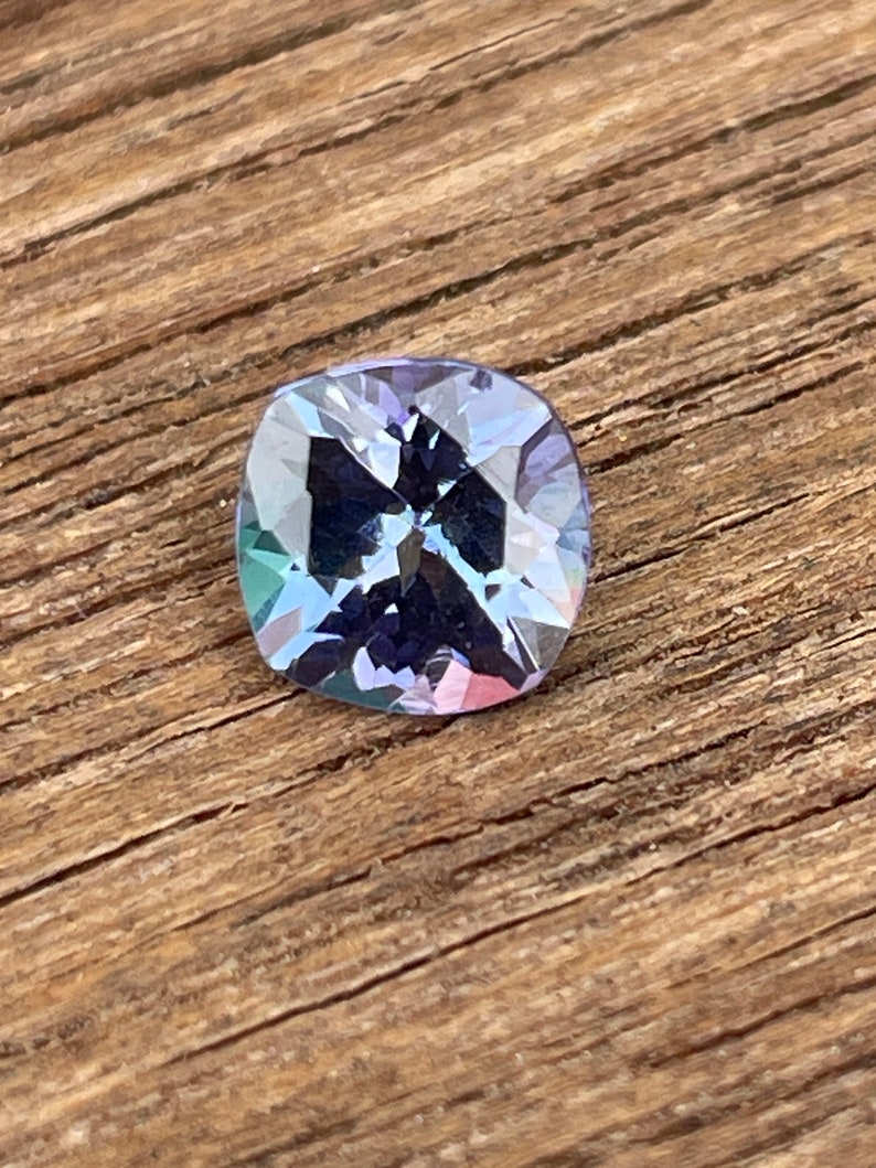 May include: A cushion-cut gemstone with a purple hue and iridescent rainbow reflections. The gem is set against a wooden background.