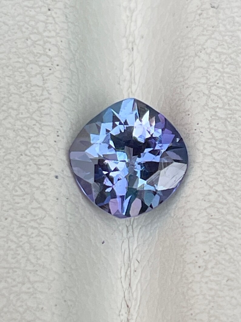 May include: A cushion-cut gemstone with a purple and blue hue. The gem is faceted and has a high polish.