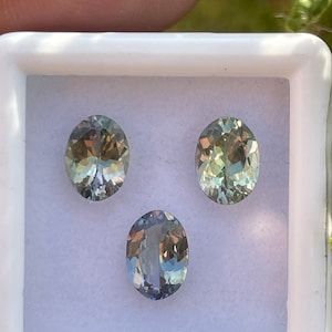 May include: Three oval-shaped gemstones in a white box. The gemstones are a light blue-green color with a slight shimmer.