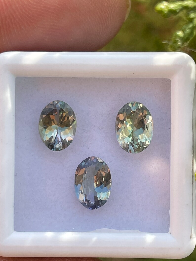 May include: Three oval-shaped gemstones in a white box. The gemstones are a light blue-green color with a slight shimmer.
