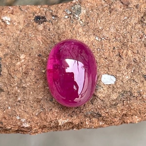 Könnte beinhalten: Ein leuchtend rosa ovaler Edelstein mit einer glatten, polierten Oberfläche. Der Edelstein ist vor einem rauen, braunen Hintergrund platziert.