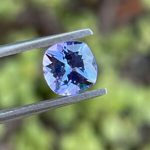 May include: A cushion-cut, purple gemstone with a slight blue hue is held by tweezers against a blurred green background.