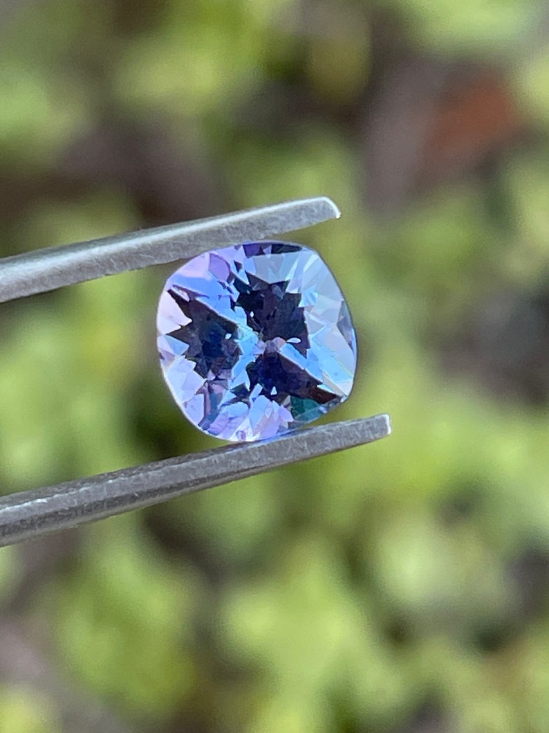 May include: A cushion-cut, purple gemstone with a slight blue hue is held by tweezers against a blurred green background.