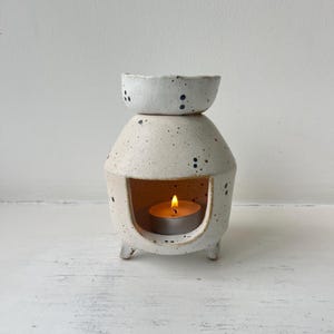 May include: A ceramic oil burner with a speckled off-white finish and dark blue spots. The structure features a wide base with three small feet, a central opening for a tea light candle, and a shallow bowl at the top for holding scented oils.