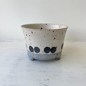 May include: A ceramic planter with a speckled off-white upper body featuring dark brown flecks and a row of black circular dots. The base is textured with horizontal ridges in a light grey finish and supported by three small, integrated feet.