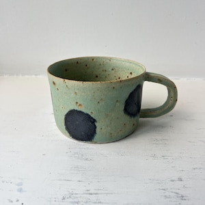 Può includere: Una tazza in ceramica fatta a mano con una glassa verde acqua macchiata. Due grandi macchie blu scuro decorano l'esterno. La tazza ha un corpo arrotondato e un piccolo manico, ideale per tè o caffè.