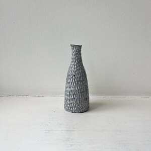 May include: A small, slender ceramic vase with a narrow neck and a wide base. The exterior surface features a textured, repeating pattern of vertical indentations in a light gray color with subtle brown speckles throughout the glaze finish.