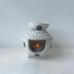 May include: A white ceramic oil burner with a speckled texture and dark gray polka dots. The piece features a wide, open front chamber containing a lit tea light candle and a small bowl at the top for wax or oil, supported by three short legs.