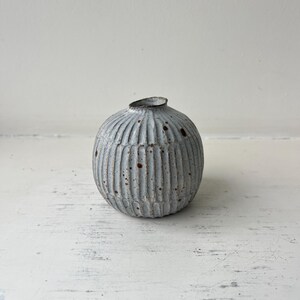 Peut inclure: Petit vase rond en céramique grise avec des détails côtelés verticaux. Le vase a une ouverture étroite et une surface texturée et tachetée. La couleur neutre et le design simple en font une pièce décorative polyvalente.