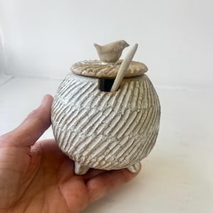 May include: A round, off-white ceramic jar with a textured, diagonal-grooved surface and three small feet. It features a matching lid topped with a small bird figurine and includes a slender ceramic spoon resting in a side notch on the rim.