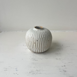 Peut inclure: Petit vase rond en céramique blanche avec un extérieur texturé et nervuré verticalement. Le vase a une ouverture étroite et un bord légèrement plus foncé. Le design est minimaliste et rustique, adapté pour afficher de petites fleurs ou comme élément décoratif.
