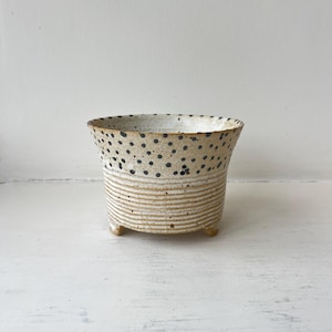 Peut inclure: Un pot en céramique avec une finition beige mouchetée, présentant des points noirs sur le bord supérieur et des stries horizontales sur la partie inférieure. Le récipient repose sur trois petits pieds et possède une large ouverture circulaire.