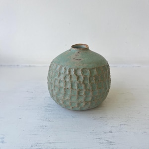May include: A small, round ceramic vase with a textured, grid-like pattern across its surface. The vase features a muted, sage-green glaze with visible brown speckles and a smooth, circular opening at the top, showcasing a rustic, earthy aesthetic.
