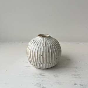 Peut inclure: Un petit vase rond en céramique blanche avec un motif texturé et nervuré. Le vase a une ouverture étroite en haut et est probablement utilisé pour exposer des fleurs ou comme objet décoratif. La couleur neutre et le design simple conviennent à divers styles d'intérieur.