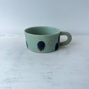 Peut inclure: Une tasse en céramique basse avec un émail vert menthe clair moucheté et des taches sombres circulaires en forme de coups de pinceau sur l'extérieur. La tasse présente une anse courte, épaisse et incurvée ainsi qu'un bord supérieur large.