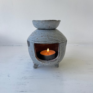May include: A speckled light blue ceramic oil burner with a rounded body, horizontal ribbed texture, and three small feet. The top features a shallow basin for oil, while the open front cavity contains a small, lit tea light candle with a flame.
