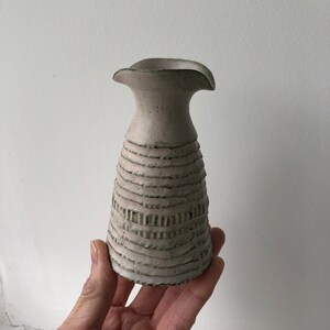May include: A handmade ceramic vase with a textured, ridged design. The vase is a light beige color with subtle green accents. The top flares out slightly, creating a unique shape. The vase is held in a hand.
