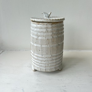 May include: A cylindrical ceramic canister with a white speckled glaze and a textured surface featuring vertical and horizontal embossed lines. The canister has a matching lid topped with a small bird figurine and stands on three short feet.
