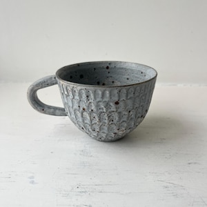 Op de afbeelding: Een kleine, grijze keramische mok met een getextureerd, gegolfd ontwerp. De mok heeft een gespikkeld glazuur en een afgeronde vorm.