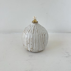 Op de afbeelding: Een witte, getextureerde keramische olielamp met een goudkleurige metalen bovenkant en een lont. De lamp heeft verticale groeven en een ronde vorm. De achtergrond is een effen wit oppervlak.