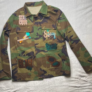 May include: A camouflage green utility jacket with various patches featuring rhinestones and sequins. The patches include the words "Stay Cute", "Stay", "Blessed", and "Glam Up".