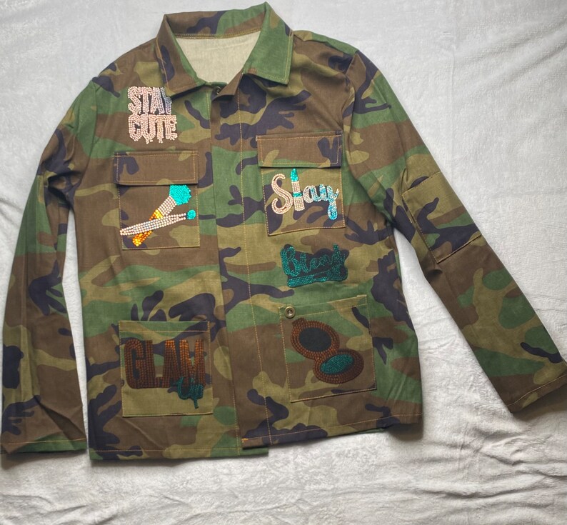 May include: A camouflage green utility jacket with various patches featuring rhinestones and sequins. The patches include the words "Stay Cute", "Stay", "Blessed", and "Glam Up".