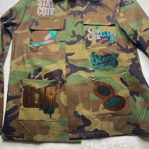 May include: A camouflage green utility jacket with various rhinestone embellishments. The jacket has multiple pockets and features the words "Stay Cute", "Stay", "Glam", and "Blend" in rhinestones.