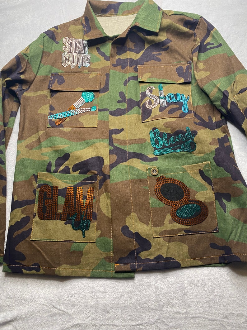 May include: A camouflage green utility jacket with various rhinestone embellishments. The jacket has multiple pockets and features the words "Stay Cute", "Stay", "Glam", and "Blend" in rhinestones.
