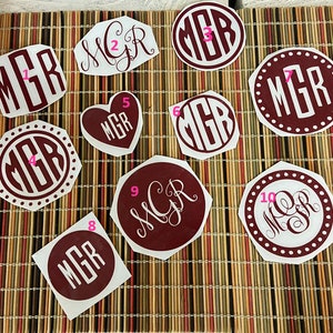 Könnte beinhalten: Zehn burgunderfarbene Monogramm-Aufkleber mit weißen Buchstaben. Die Monogramme haben verschiedene Formen, darunter Kreise, Ovale, Herzen und Achtecke. Die Buchstaben "MGR" sind auf jedem Aufkleber zu sehen.