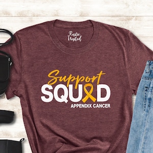 May include: Burgundy t-shirt with white text that reads "Support Squad" with a yellow ribbon that says "Appendix Cancer".