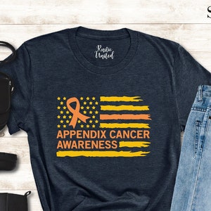 May include: A navy blue t-shirt with an orange and yellow flag design and the words "APPENDIX CANCER AWARENESS". The image also includes black boots, a black purse, blue jeans, and sunglasses.