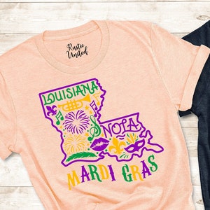 May include: Two t-shirts with a graphic design of the state of Louisiana with the words "Louisiana" and "Mardi Gras" printed on them. The shirts are in peach and dark blue.