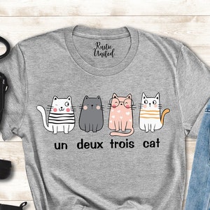 以下が含まれることがあります： 4匹の猫が並んだデザインのグレーのTシャツ。猫はそれぞれ違う服を着ていて、表情も違います。「un deux trois cat」という文字が猫の下に印刷されています。