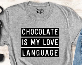 May include: A gray t-shirt with the text "CHOCOLATE IS MY LOVE LANGUAGE" in a black and white design. The image also includes a black purse, black boots, and blue jeans with sunglasses.