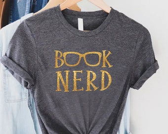 May include: Dark gray t-shirt with rolled-up sleeves, featuring the text "BOOK NERD" in gold glitter. The shirt is hanging on a wooden hanger with a light blue button-down shirt, a pair of jeans, and a brown hat in the background.