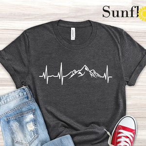 May include: Dark grey heather t-shirt with a white mountain range and heartbeat graphic. The shirt is laid out on a wooden surface with a pair of red sneakers and blue jeans.