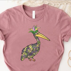May include: A mauve t-shirt with a cartoon pelican wearing a green, purple, and gold jester hat. The pelican has a green, purple, and gold camouflage pattern.