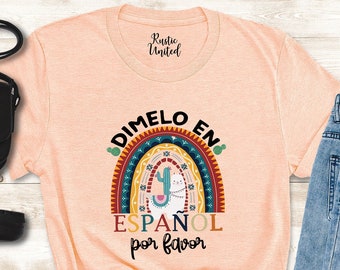 May include: A peach-colored t-shirt with a rainbow graphic and the text "Dimelo en Espanol por favor". The shirt is paired with blue jeans, black ankle boots, and a black crossbody bag.