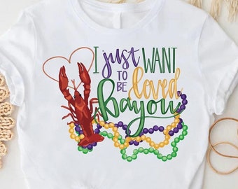 May include: White t-shirt with a purple, green, and gold Mardi Gras beads design. The shirt also features a red crawfish and the text "I just want to be loved bayou".
