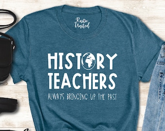 May include: Teal blue t-shirt with white text that reads "History Teachers Always Bringing Up The Past". A small globe graphic is above the word "Teachers". The shirt is laid out on a woodgrain surface with a black purse, black boots, and blue jeans.