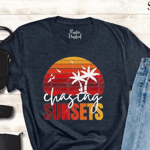 May include: A navy blue t-shirt with a vintage sunset graphic and the text "chasing sunsets". The graphic features palm trees and birds flying over a red, orange, and yellow sunset.