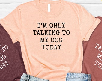 May include: Three t-shirts with the text "I'm only talking to my dog today" in white lettering on a maroon, peach, and heather gray background. The shirts are laid out on a wooden surface with denim shorts and sandals.